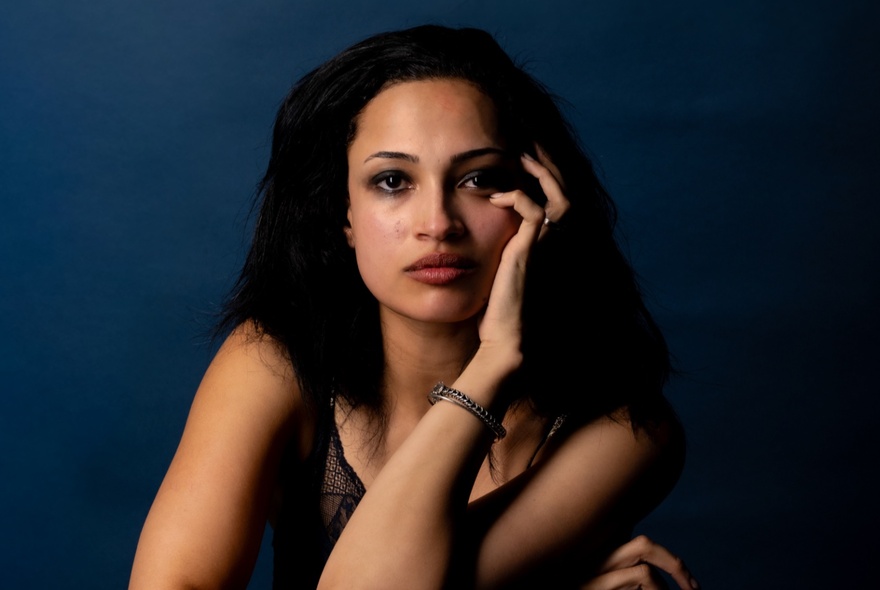Actor Sarah Ruhl posing with her hand on her face and looking blankly ahead in a dark navy room. 