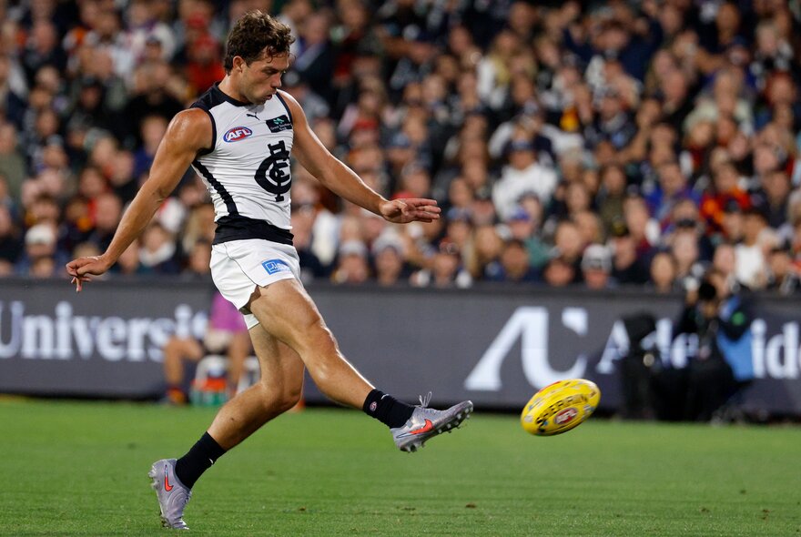 A Carlton AFL football player with his leg raised in the air, having just kicked the football during a match, a blurred stadium crowd in the background.