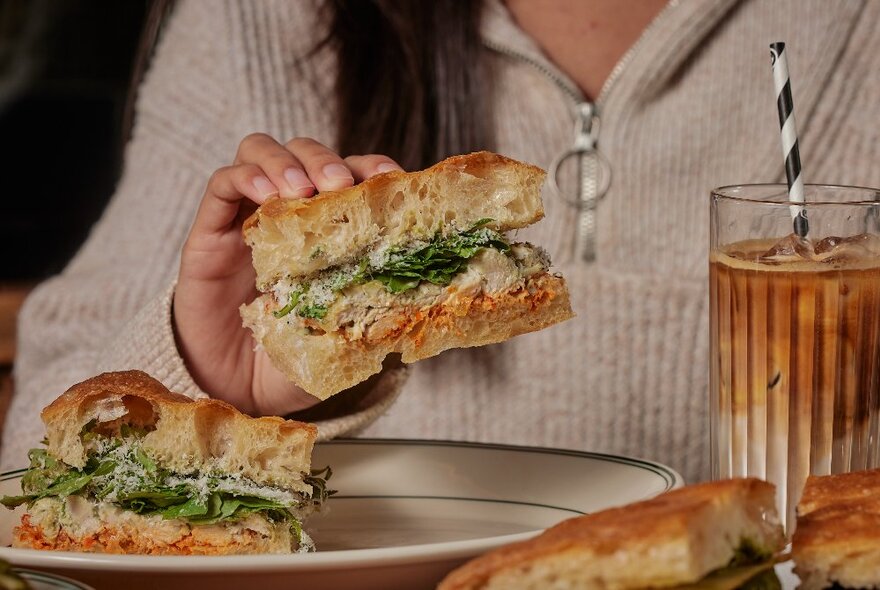 Hands holding large focaccia sandwich over a plate with a tall drink on the side.