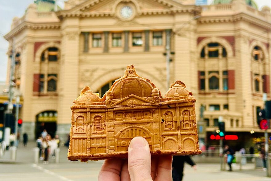 A hand holding up a baked flat waffle shaped like Flinders Street Station, with the real Flinders Street Station in the background.