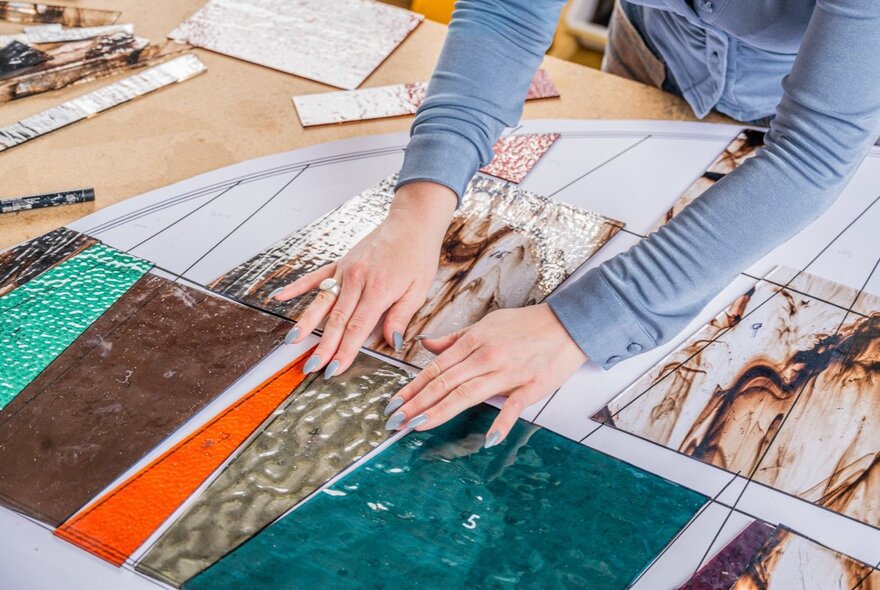 A person arranges various pieces of coloured, textured glass on a table.
