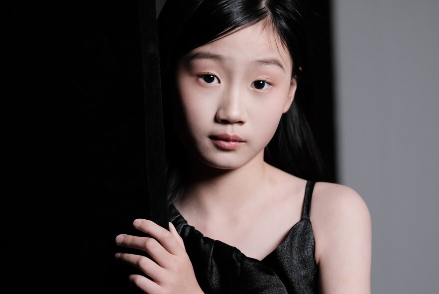 A young teenager, with straight black hair and wearing a black dress, leaning against a dark wall.