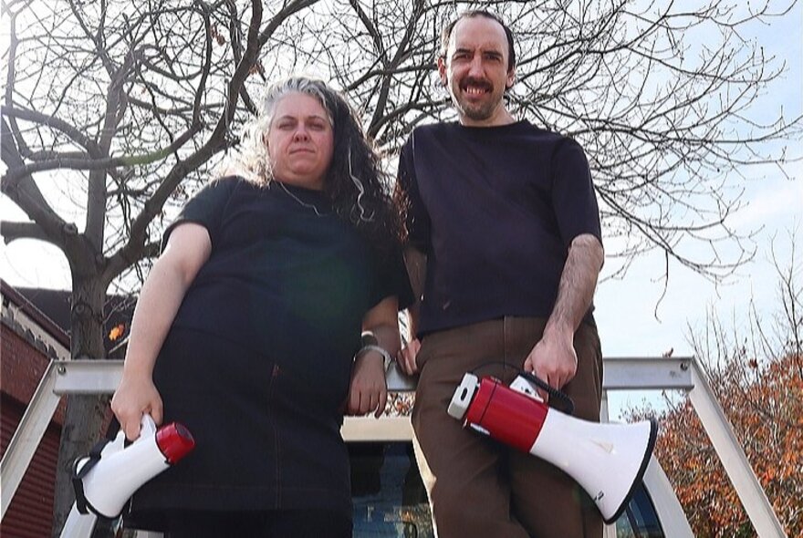 Two actors holding megaphones in front of bare trees.