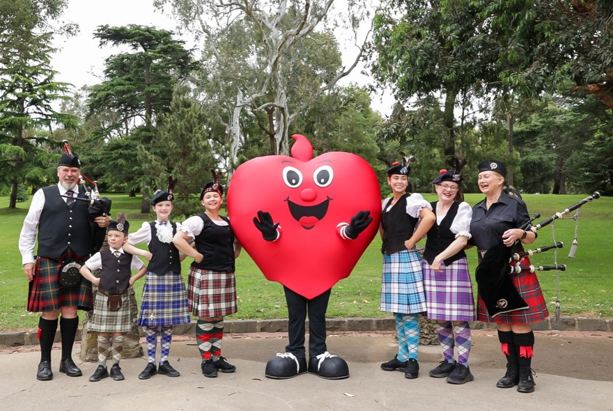A line of adults and children wearing kilts, two of them holding bagpipes, posing for a photo in the Kings Domain Gardens with a red smiling heart-shaped mascot.