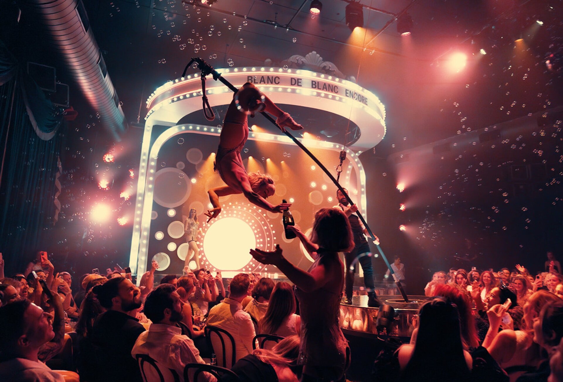 A cabaret show with a performer balancing on pole about to pour a bottle of champagne