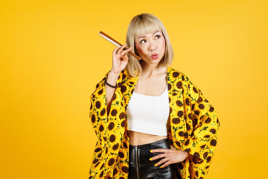 Japanese comedian Showko in a yellow sunflower print jacket against the same yellow shaded background looking upwards. 
