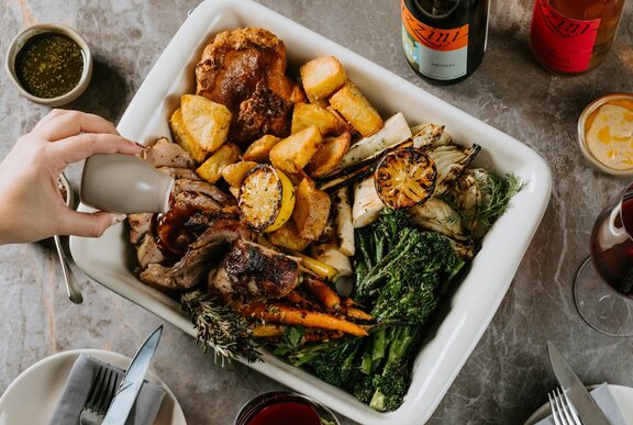 A white roasting tray filled with roast vegetables alongside a quartered roast chicken.