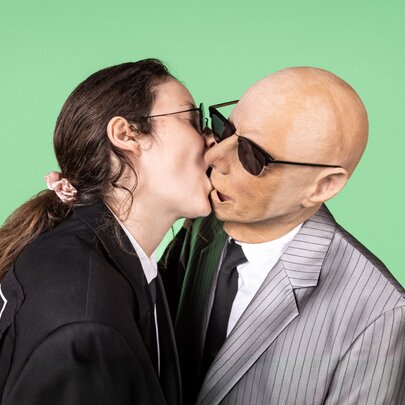 A person with long brown hair tied back, wearing a black blazer and sunglasses, leans forward to kiss a person wearing a rubber bald head face mask, in a grey pinstripe suit and sunglasses, set against a solid bright green background.