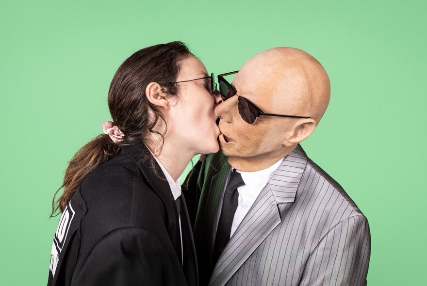 A person with long brown hair tied back, wearing a black blazer and sunglasses, leans forward to kiss a person wearing a rubber bald head face mask, in a grey pinstripe suit and sunglasses, set against a solid bright green background.