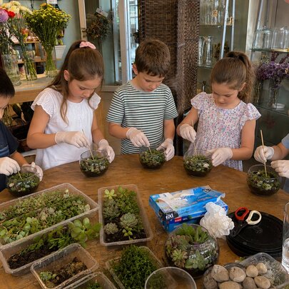 A group of children wearing protective gloves while participating in a hands-on, educational terrarium-building workshop around a wooden table.