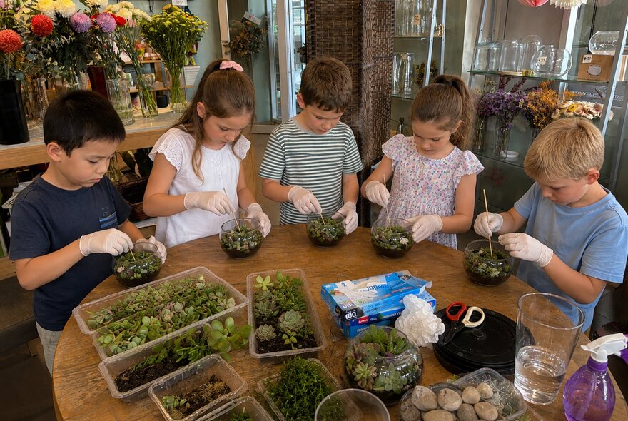A group of children wearing protective gloves while participating in a hands-on, educational terrarium-building workshop around a wooden table.
