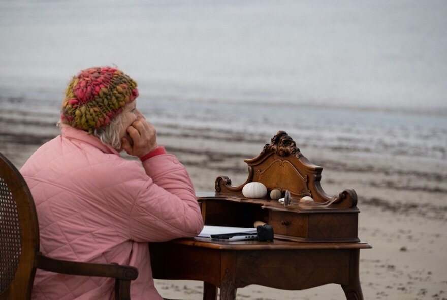 A woman wearing a pink jacket and beanie, seated at an antique writers desk on a seaweed-strewn beach.