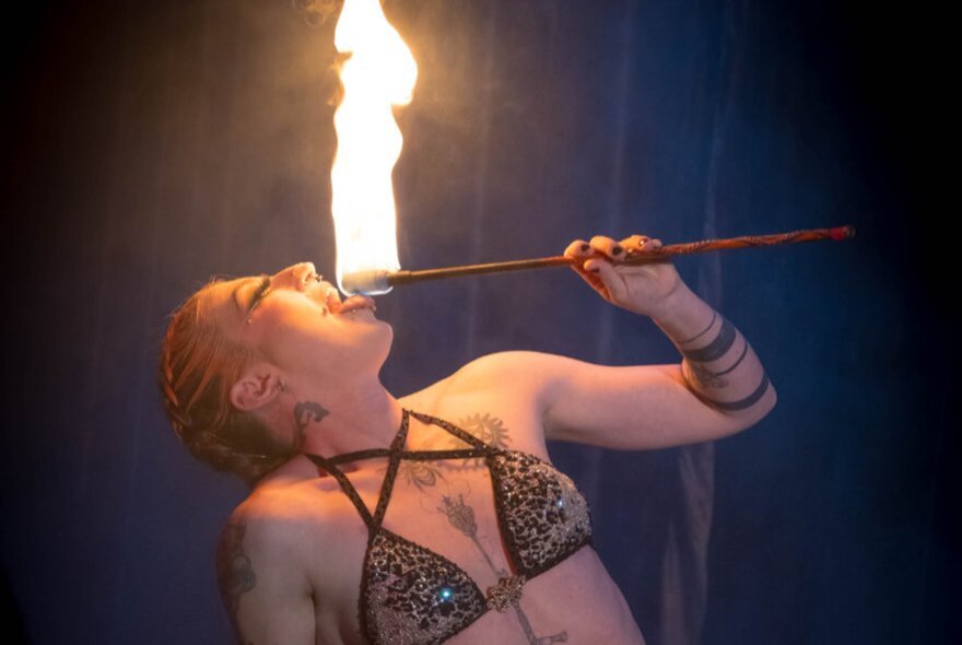 A performer wearing a bikini and holding a firestick above her open mouth in a small cabaret venue.