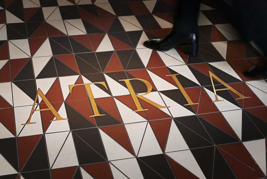 A striking tiled floor with the word Atria in gold and a pair of women's black heeled boots walking through. 