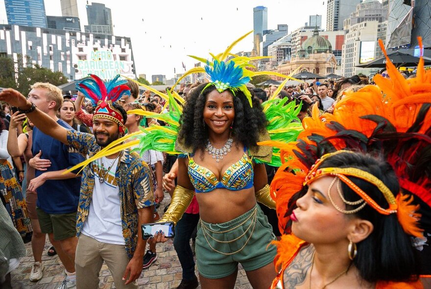 A crowd of people enjoying themselves at Fed Square at an outdoor daytime festival event with dancers in costumes in the crowd.