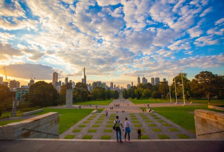 Two people standing on the edge of the Shrine, looking out over concrete and grass squares with Melbourne CBD buildings in the background.