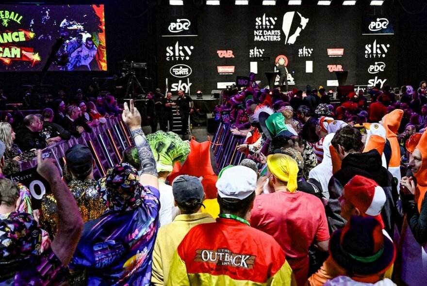 A large crowd of people in colourful fancy dress costumes packed closely together at an indoor darts tournament.