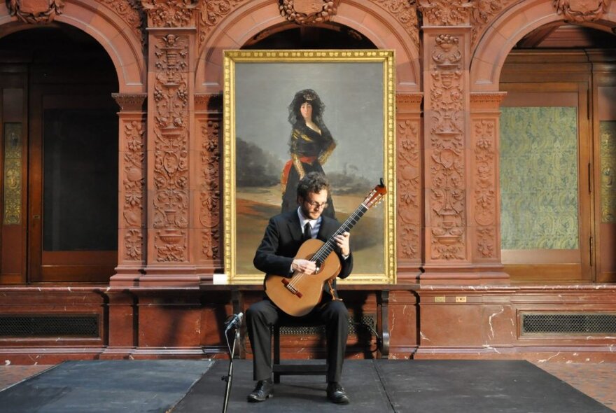 Seated man playing a classical guitar in front of a large oil painting.