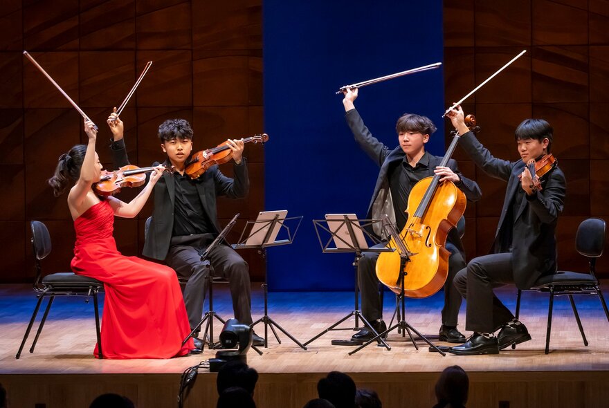 Four young members of a chamber music group performing on stage with batons in the air. 