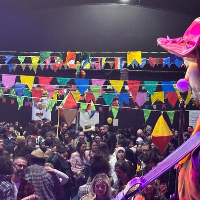 A traditional Brazilian musician on stage in colourful costume in front of a large crowd seated underneath brightly coloured bunting.