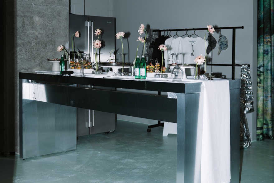 A studio or a workshop, featuring a long table with multiple floral arrangements in vases.