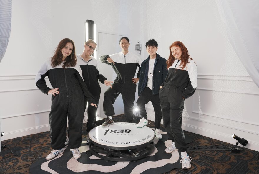 A group of five people in black and white tracksuits, posing around a white disc on the floor or a white room. 