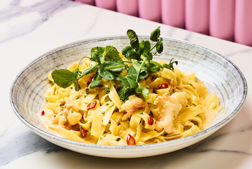 A bowl of tagliatelle pasta topped with garnish on a marble table.