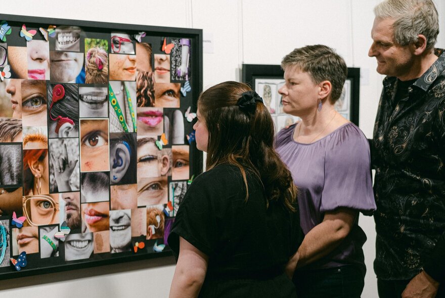 Three people looking at a large framed artwork made up of images of faces.