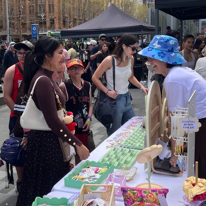 People at an outdoor market interacting with stallholder wearing a blue hat. 