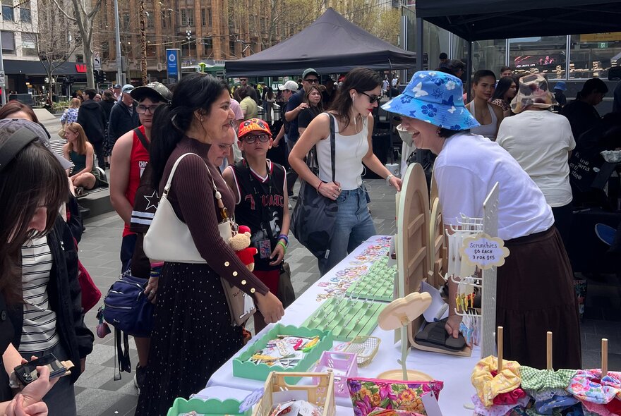 People at an outdoor market interacting with stallholder wearing a blue hat. 