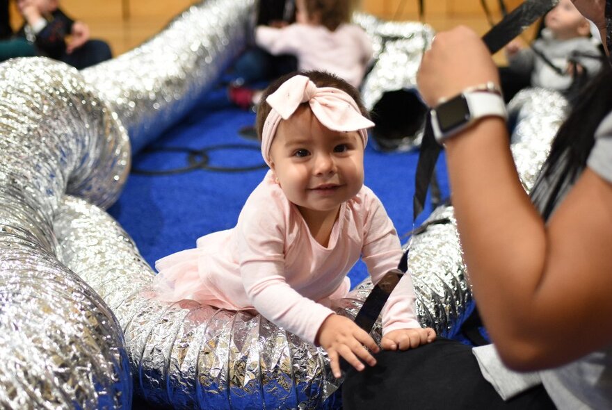 A smiling tot with a large bow in around her head playing in a room with flexible silver piping. 