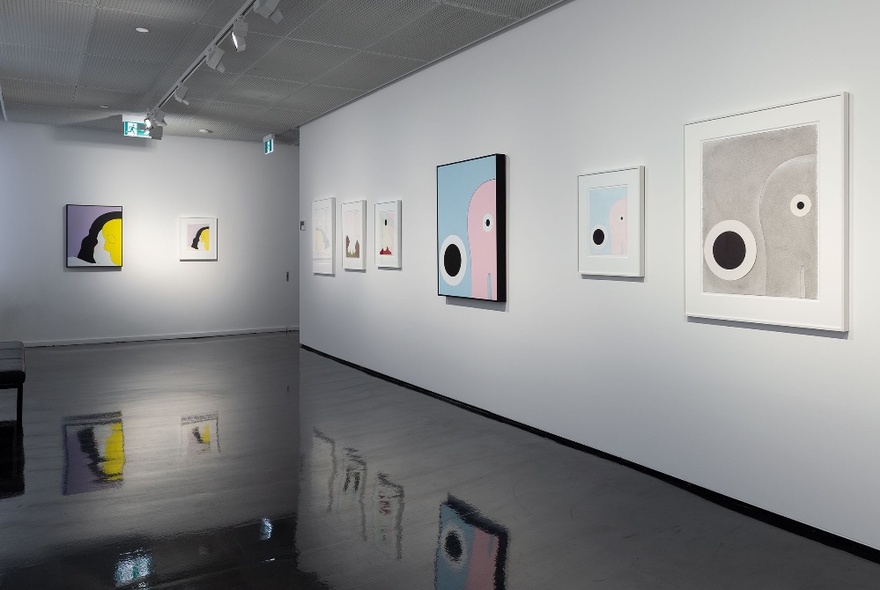 A series of framed paintings and drawings hanging on a white gallery wall, all spotlit.