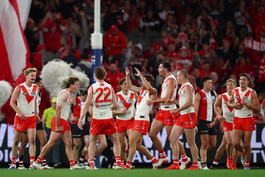 A large huddle of Sydney Swans  AFL players celebrating on the field in front of the goal square, with blurred fans in the stadium in the background.