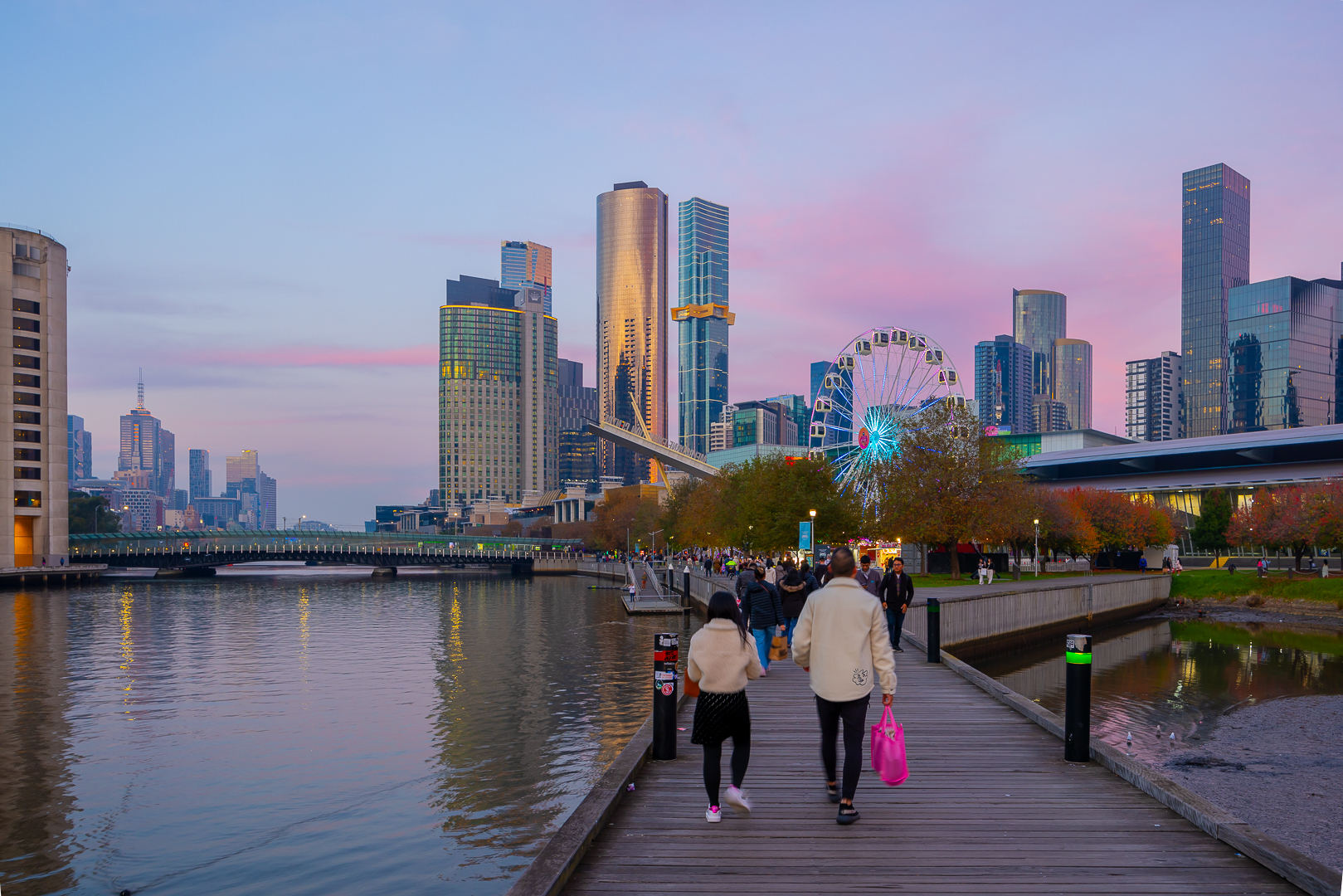 Crowds walking towards a waterside Ferris wheel in the city at sunset.