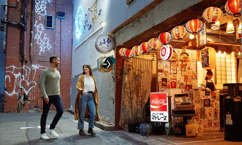 Two friends are walking into a small restaurant in a laneway. There are glowing lanterns hanging outside.