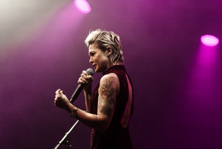 A singer in profile, singing into a microphone she holds in her hands, on a stage.