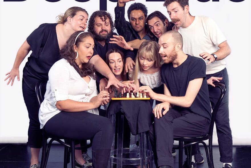 Performers on a stage, all sitting or standing around and reaching out to small centre table that features a chess set on it.