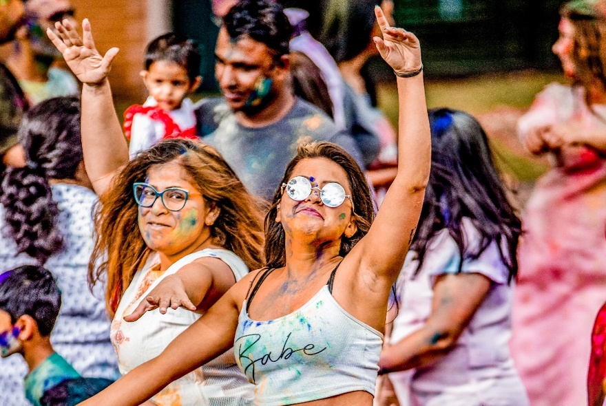 Two people in a crowd celebrating and dancing, covered in coloured powder, at Melbourne's outdoor Holi Festival.