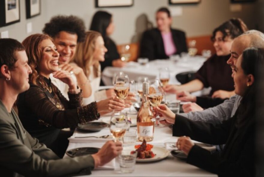 People seated around a table, eating, drinking, and smiling, with plates of food and glasses of wine on the table.