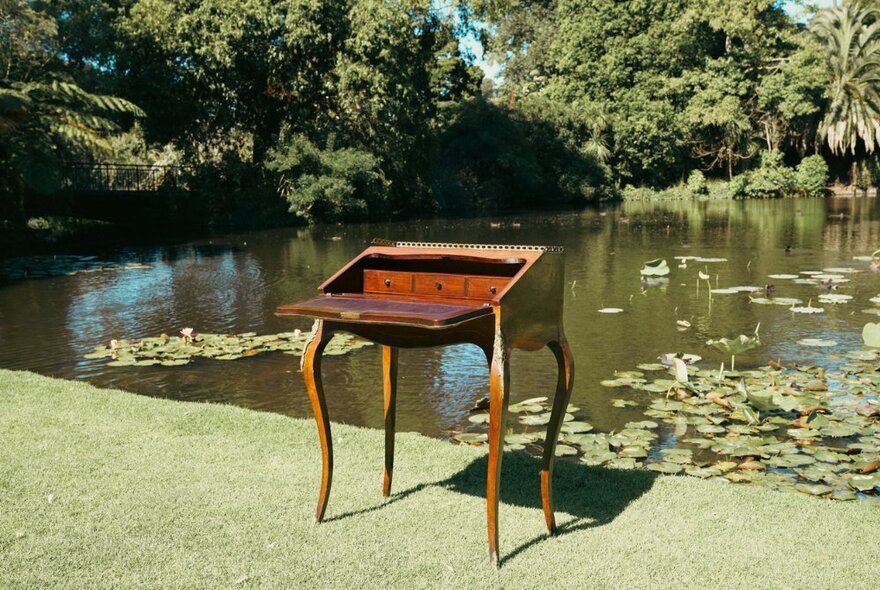 An antique writers desk on the grass beside a lily pond with trees.