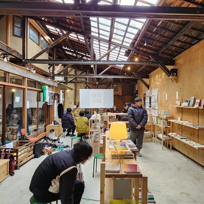 The interior of a ply-lined warehouse with artworks displayed on ply shelves and benches and people walking around.