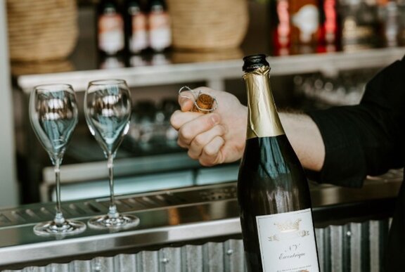 Two empty wine glasses on a steel bar beside a person's arm holding the cork of a wine bottle in the foreground.