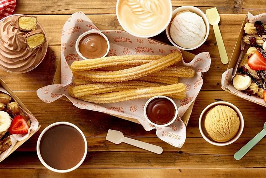 Wooden table with central platter of churros and chocolate dips, with drinks, cups and spoons.