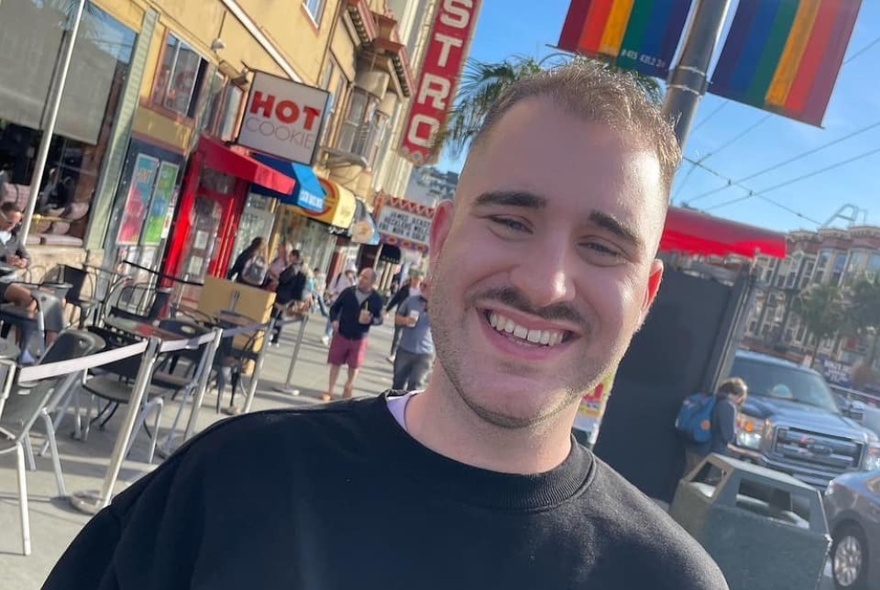 Portrait of a smiling man wearing a black t-shirt on the footpath near a cafe with outdoor seating.