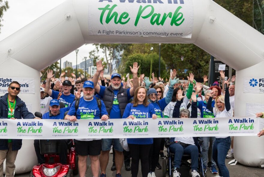 A crowd of people in blue t-shirts waiting to start a charity walk behind a white ribbon, underneath a white blow-up archway.