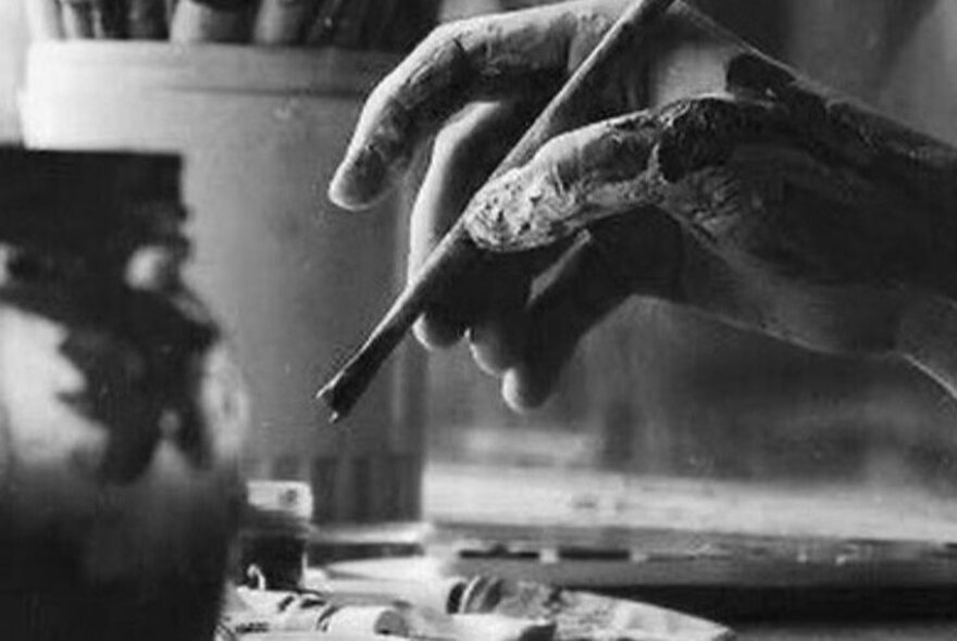 A black and white photograph of an artist's paint-covered hand holding a blunt-ended paint brush.