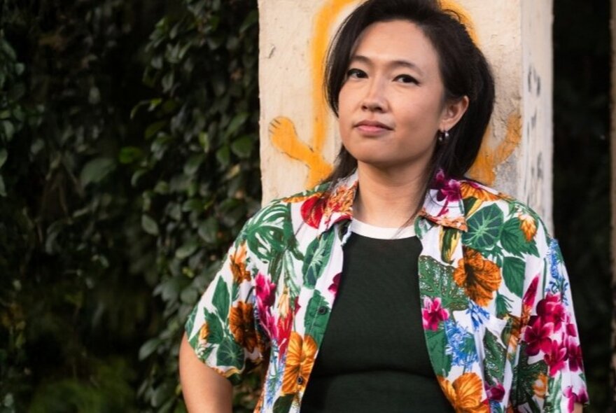 Portrait of a Singaporean woman wearing a brightly coloured floral shirt, standing in front of a concrete pole with yellow graffiti on it.
