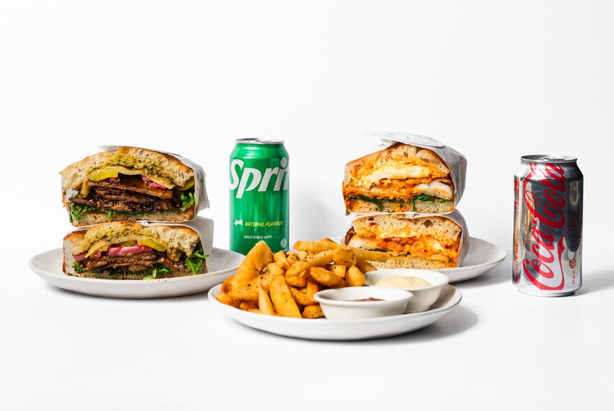 Two sandwiches on white plates, a bowl of chips and a can of Sprite and Coke.