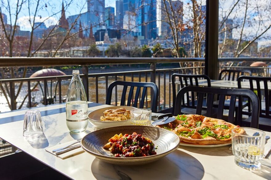 An outdoor restaurant by the river with pizza and pasta dishes on the table.