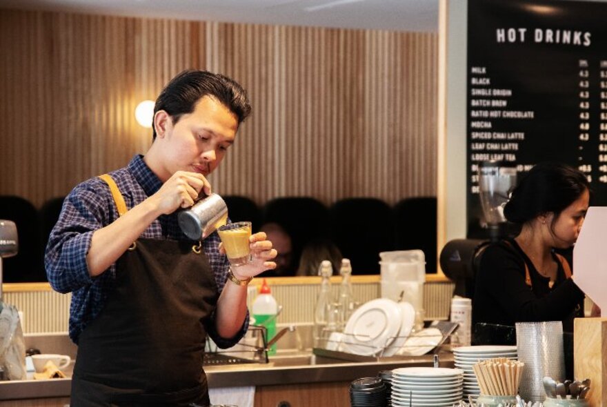 A barista pouring milk into a glass of coffee, with a menu of hot drinks on the wall to his right.
